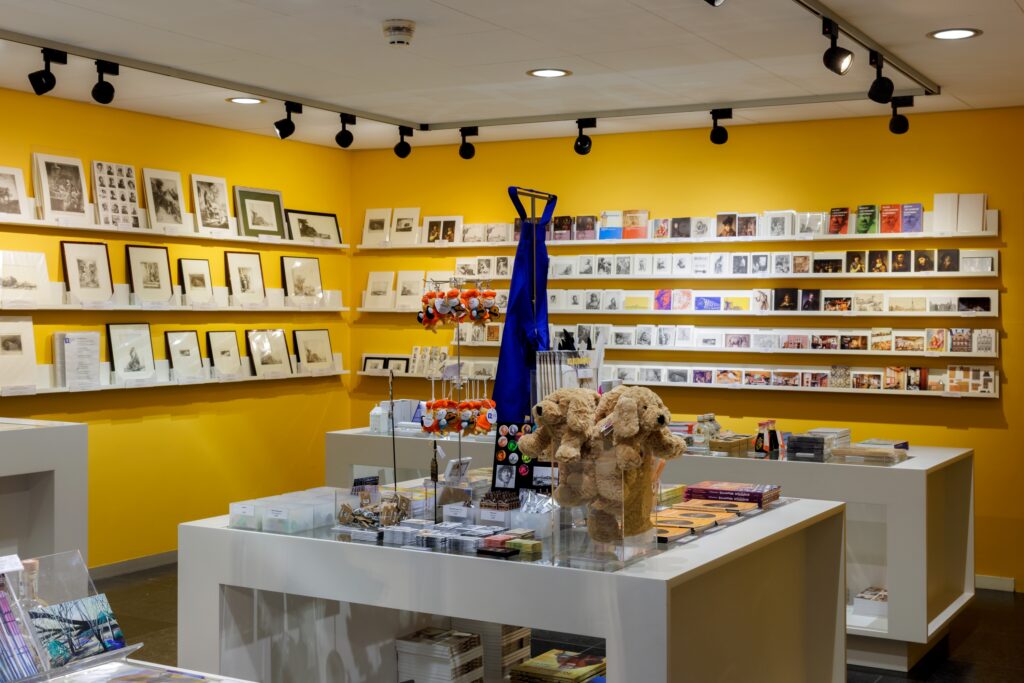 De museumwinkel van Museum Rembrandthuis met een gele wand vol met ansichtkaartjes, de knuffel Kwast, en andere items in de winkel